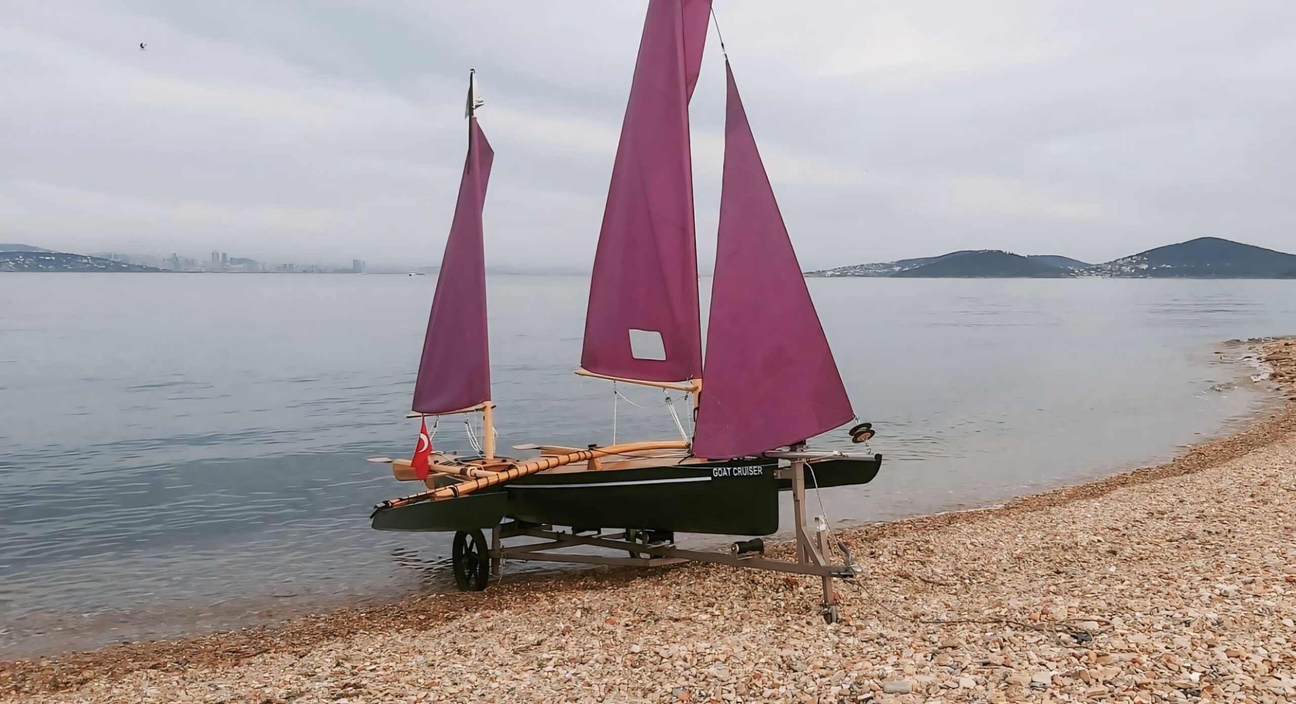 The plywood Goat Cruiser trimaran on the shore with its pink sails hoisted