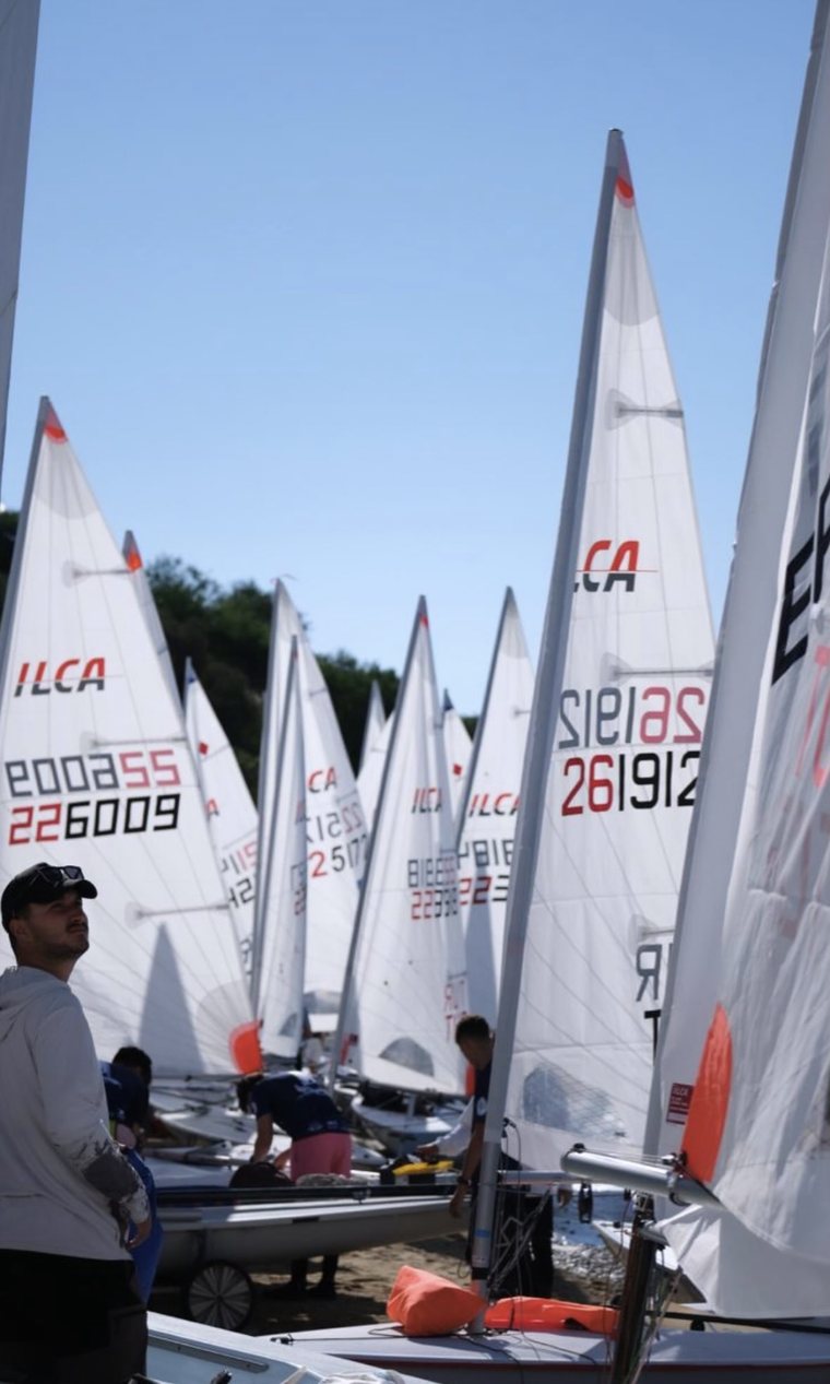 Kumsalda yan yana ILCA yelkenleri ve yarışa hazırlanan sporcular